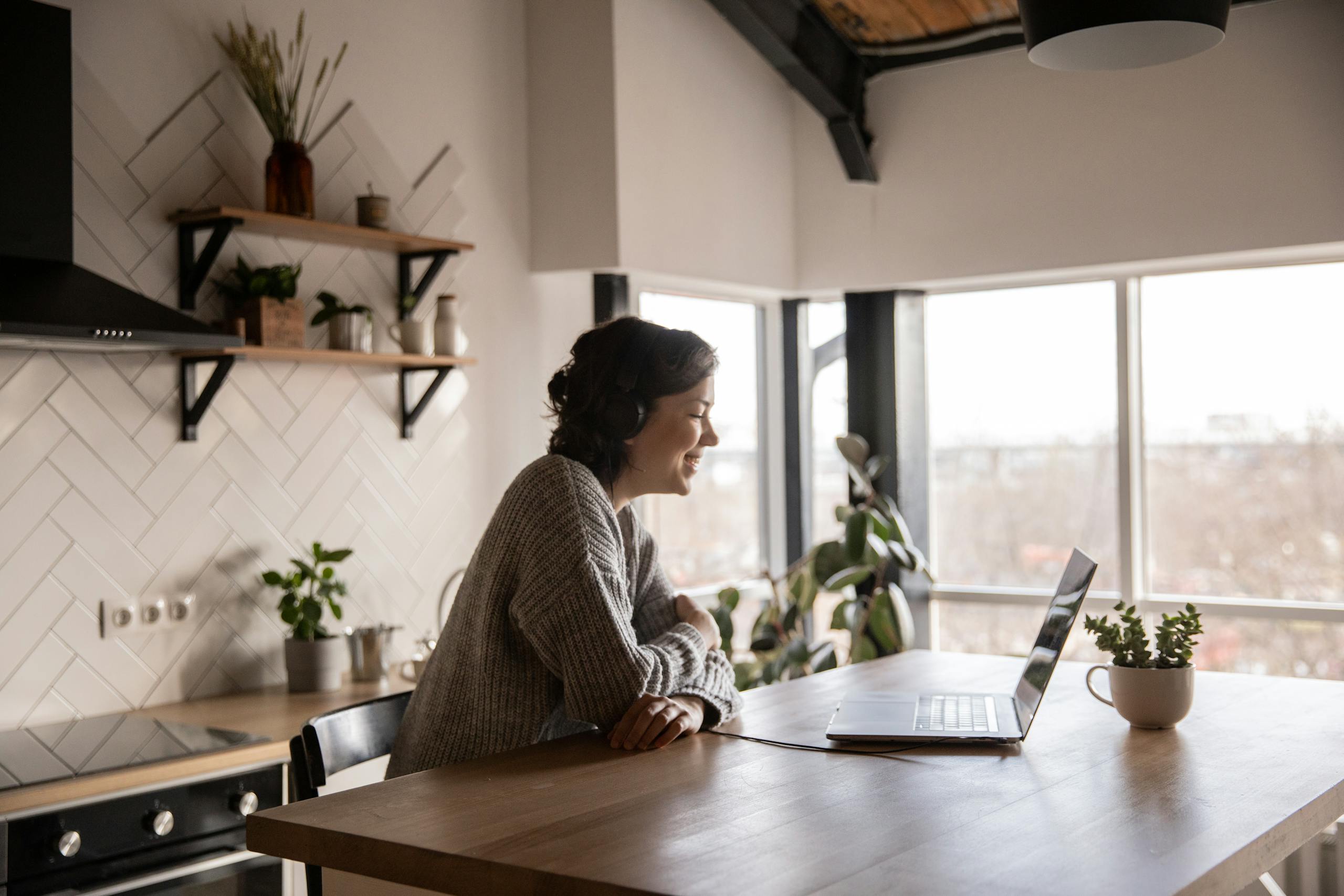 A woman enjoys a video call on her laptop in a modern, cozy kitchen setting with natural light.
