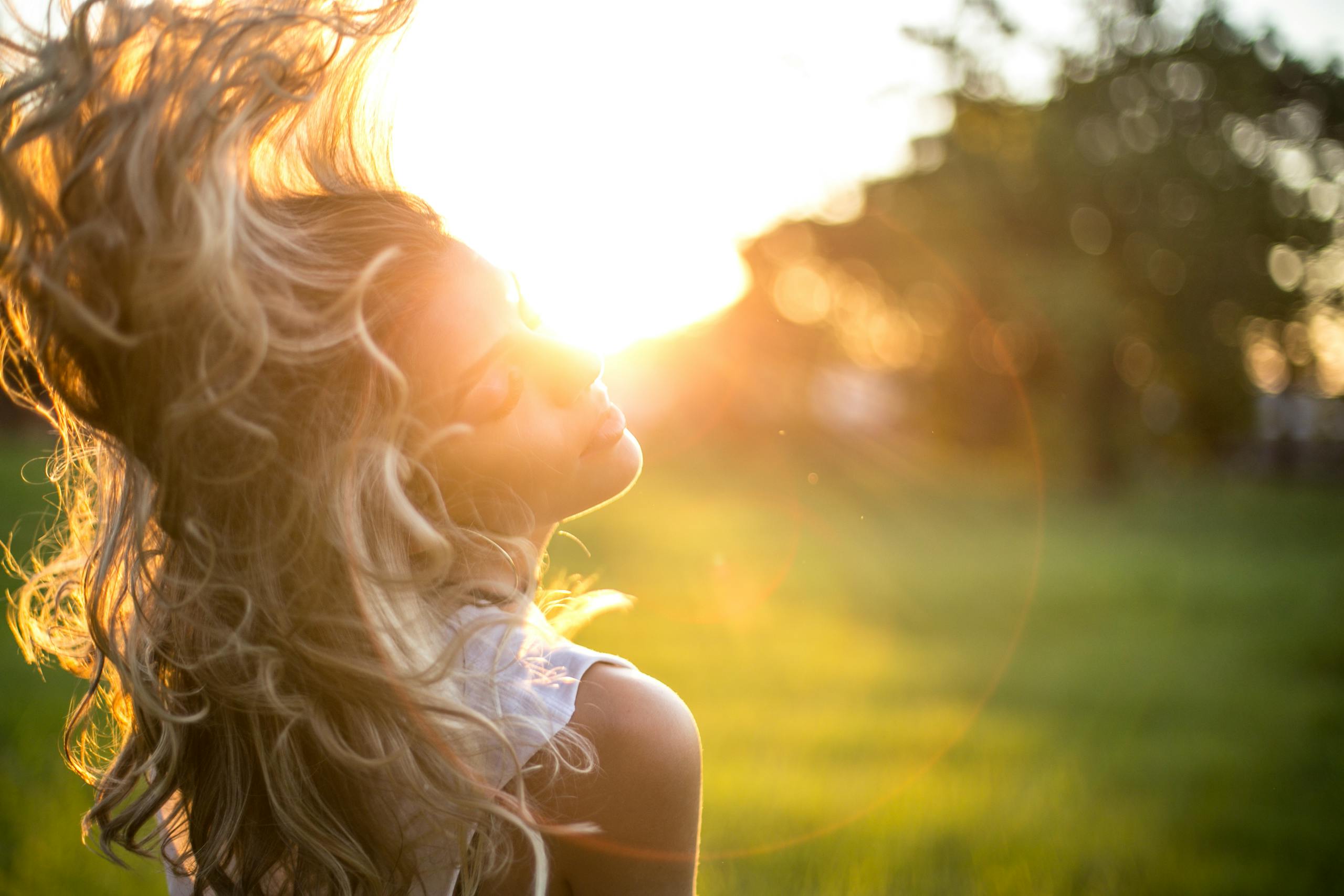 A young woman tosses her long, wavy blond hair in the sunlight during golden hour outdoors.