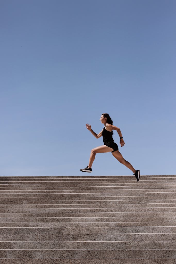 Athletic woman sprinting up outdoor stairs under clear blue sky, showcasing fitness and strength.