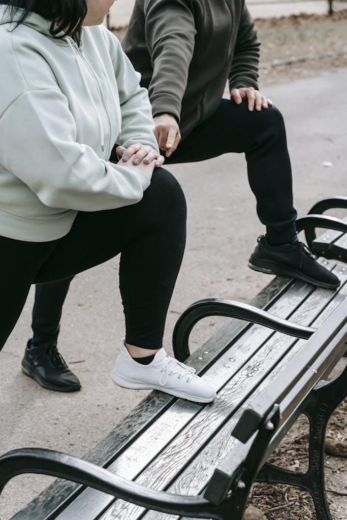 Crop anonymous trainer and overweight woman doing exercises on wooden bench in park in daytime